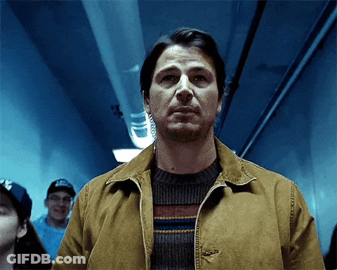 A Man In A Brown Jacket And Sweater Stands In A Hallway GIF