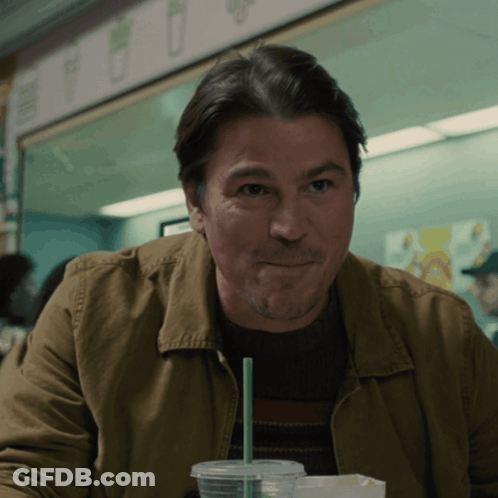A Man In A Brown Jacket Is Sitting At A Table With A Cup Of Coffee And A Green Straw GIF