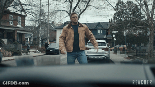 A Man In A Brown Jacket Is Walking Down A Street With The Word Reacher On The Bottom Right GIF