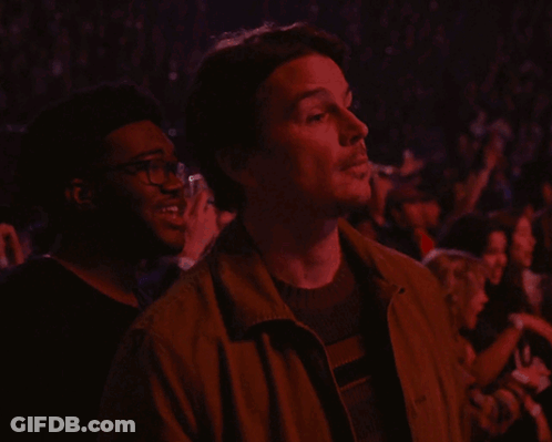 A Man In A Brown Jacket Looks Up At Something In A Crowd GIF