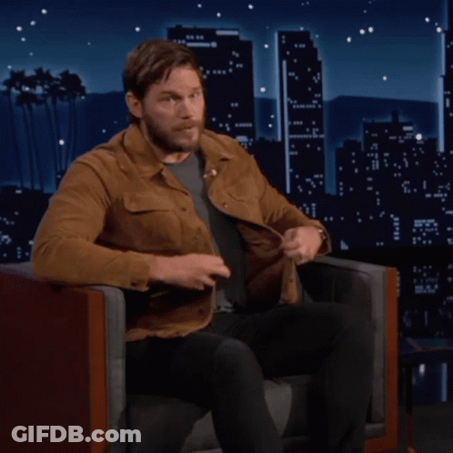 A Man In A Brown Jacket Sits In A Chair In Front Of A City Skyline GIF