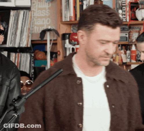 A Man In A Brown Jacket Stands In Front Of A Sign That Says ' Philip Glass Piano Studio ' GIF
