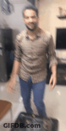 A Man In A Brown Shirt And Blue Jeans Is Standing In A Living Room . GIF