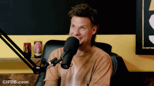 A Man In A Brown Shirt Is Talking Into A Microphone With A Can Of Beer On A Shelf Behind Him GIF