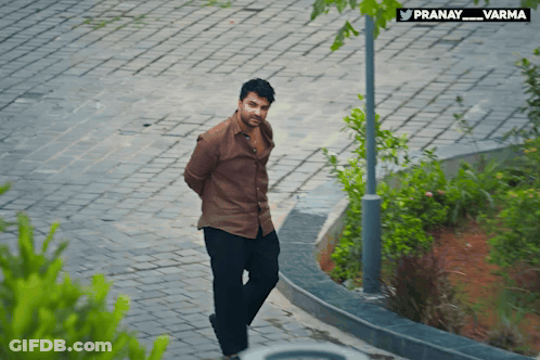 A Man In A Brown Shirt Is Walking Down A Brick Sidewalk With A Sign That Says Pranay_varma GIF