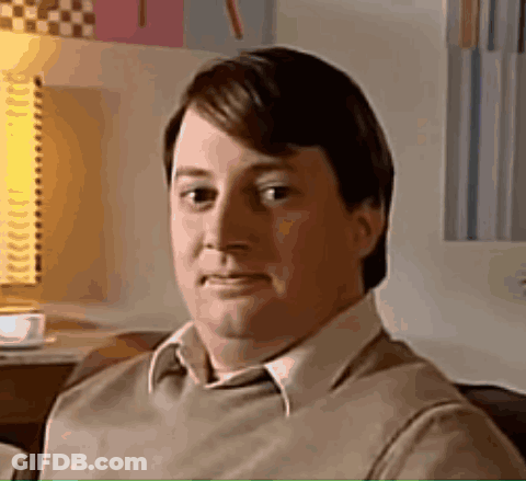A Man In A Brown Sweater And White Shirt Is Sitting On A Couch Looking At The Camera . GIF