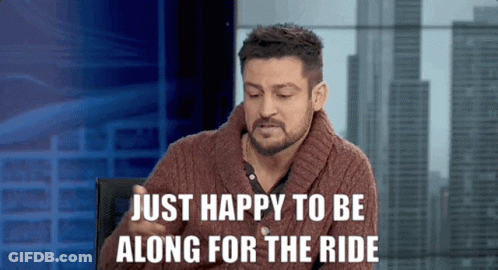 A Man In A Brown Sweater Is Sitting In Front Of A Sign That Says Just Happy To Be Along For The Ride GIF