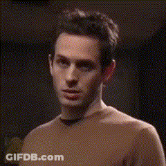 A Man In A Brown Sweater Is Standing In A Dark Room Looking At The Camera . GIF