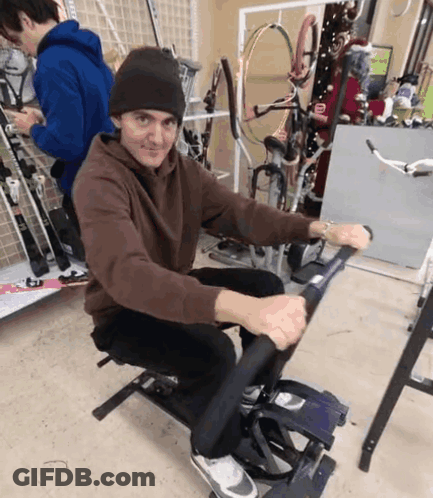 A Man In A Brown Sweatshirt Is Sitting On A Machine GIF