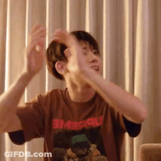 A Man In A Brown T-Shirt Is Sitting On A Couch With His Arms Outstretched In Front Of Curtains . GIF