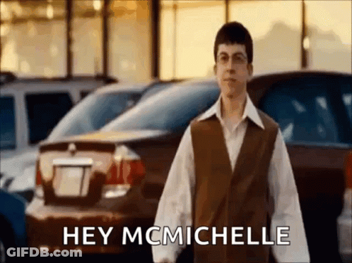A Man In A Brown Vest Is Standing In Front Of A Parking Lot And Says Hey Mcmichelle GIF
