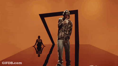A Man In A Camo Hoodie Stands In Front Of A Black Triangle GIF