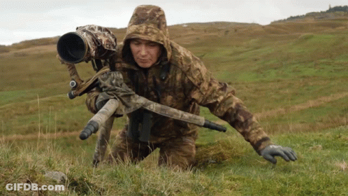 A Man In A Camo Jacket Is Kneeling Down In The Grass With A Camera On A Tripod GIF