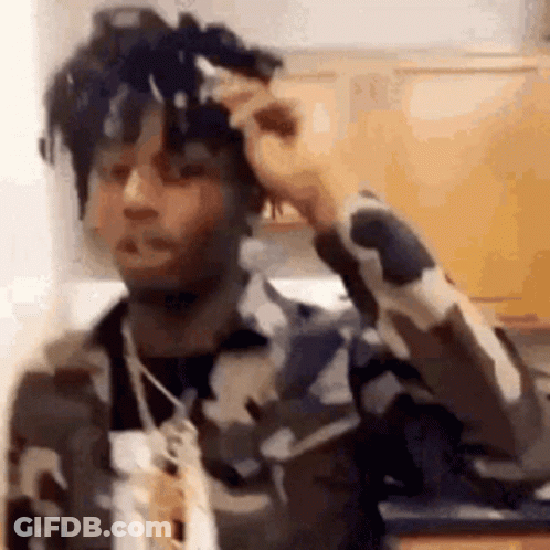 A Man In A Camouflage Jacket Is Standing In A Kitchen With His Hand On His Hair . GIF