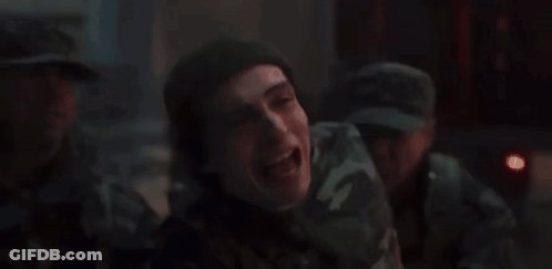 A Man In A Camouflage Uniform Is Laughing In A Dark Room With Other Men . GIF
