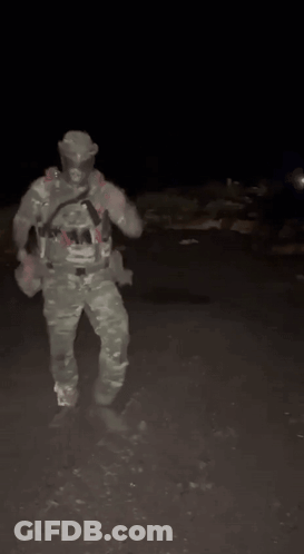 A Man In A Camouflage Uniform Is Standing In The Dark Holding A Gun . GIF