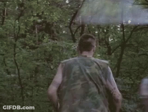 A Man In A Camouflage Vest Is Running Through The Woods With A Smoke Bomb . GIF