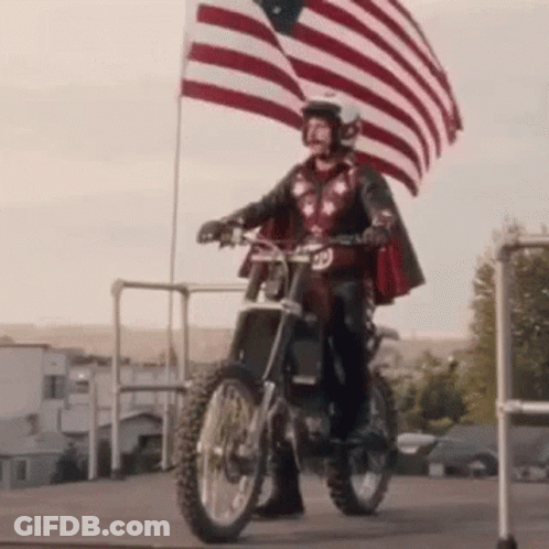 A Man In A Cape Is Riding A Dirt Bike While Holding An American Flag . GIF