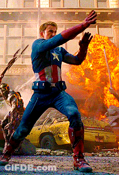 A Man In A Captain America Costume Is Standing In Front Of A Huge Explosion GIF
