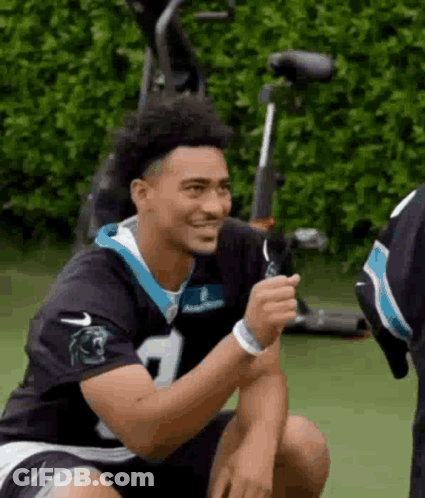 A Man In A Carolina Panthers Jersey Is Sitting On The Ground And Smiling . GIF