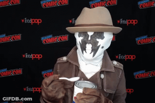 A Man In A Costume Is Holding A Can Of Food In Front Of A Wall That Says Comic Con On It GIF