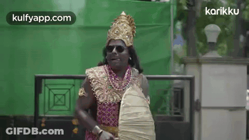 A Man In A Costume Is Standing In Front Of A Green Wall Holding A Fan And A Shield . GIF
