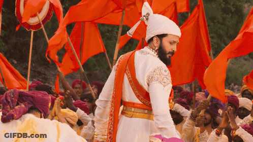 A Man In A Costume With A Beard Stands In Front Of A Crowd Of People Holding Orange Flags GIF