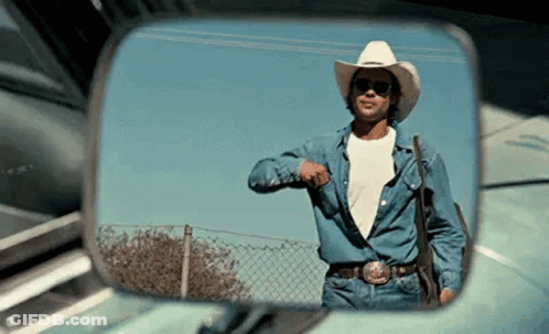 A Man In A Cowboy Hat And Sunglasses Is Standing In Front Of A Car In A Rear View Mirror . GIF