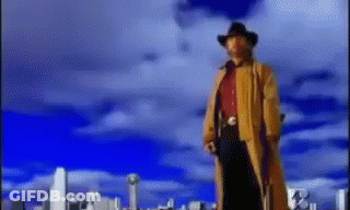 A Man In A Cowboy Hat And Trench Coat Stands In Front Of A City Skyline GIF
