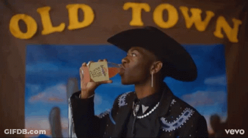 A Man In A Cowboy Hat Is Holding A Drink In Front Of An Old Town Poster GIF