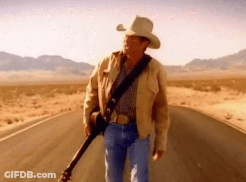 A Man In A Cowboy Hat Is Holding A Guitar While Walking Down A Road . GIF