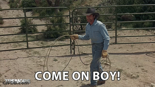 A Man In A Cowboy Hat Is Holding A Lasso In Front Of A Fence That Says Come On Boy GIF