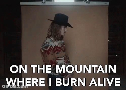 A Man In A Cowboy Hat Is Standing In Front Of A Brown Backdrop With The Words On The Mountain Where I Burn Alive GIF