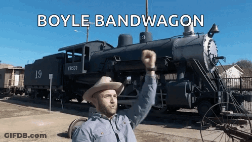 A Man In A Cowboy Hat Is Standing In Front Of A Train That Says Boyle Bandwagon On It GIF