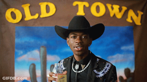 A Man In A Cowboy Hat Is Standing In Front Of An Old Town Sign GIF