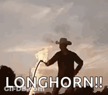 A Man In A Cowboy Hat Is Standing Next To A Fire With The Words `` Longhorn '' Written On It . GIF