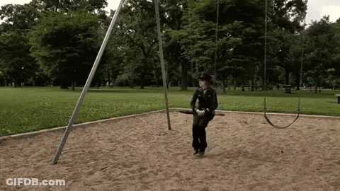 A Man In A Cowboy Hat Is Swinging On A Swing Set In A Park GIF