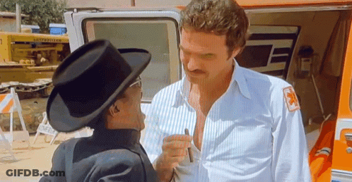 A Man In A Cowboy Hat Is Talking To Another Man In A White Shirt With A Red Cross On His Sleeve . GIF