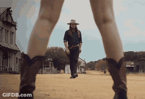 A Man In A Cowboy Hat Is Walking Down A Dirt Road Next To A Woman In Cowboy Boots GIF