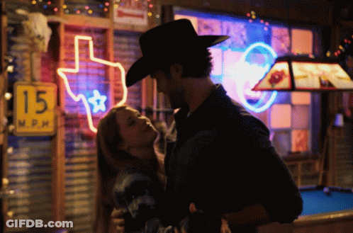 A Man In A Cowboy Hat Kisses A Woman In Front Of A Neon Sign That Says 15 Mph GIF