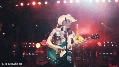 A Man In A Cowboy Hat Playing A Guitar On A Stage GIF