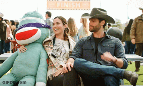 A Man In A Cowboy Hat Sits Next To A Woman Holding A Sock Monkey Under A Finish Sign GIF