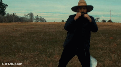 A Man In A Cowboy Hat Stands In A Grassy Field GIF