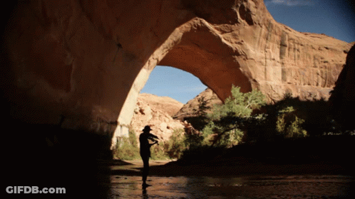A Man In A Cowboy Hat Stands Under A Large Rock Archway GIF