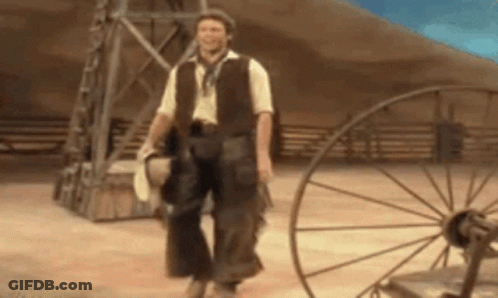 A Man In A Cowboy Outfit Is Walking In A Dirt Field . GIF