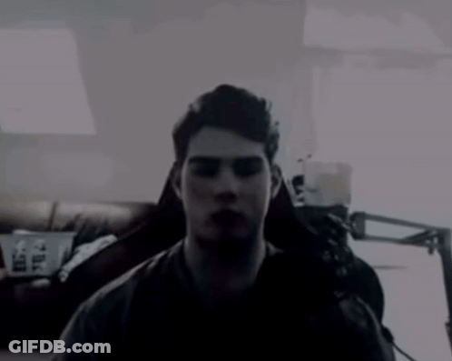 A Man In A Dark Room With A Couch And A Lamp In The Background GIF
