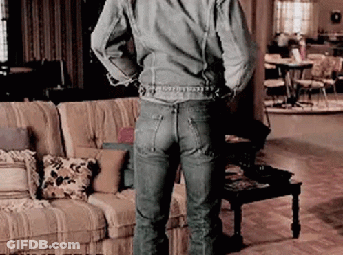A Man In A Denim Jacket And Jeans Is Standing In A Living Room With His Hands Behind His Back . GIF