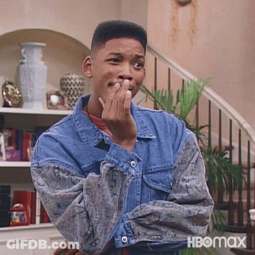 A Man In A Denim Jacket Is Covering His Mouth With His Hand While Standing In Front Of A Staircase . GIF