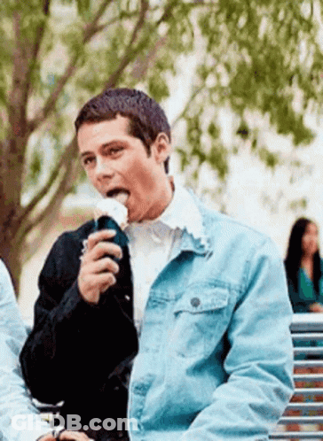 A Man In A Denim Jacket Is Sitting On A Park Bench Eating A Sandwich . GIF