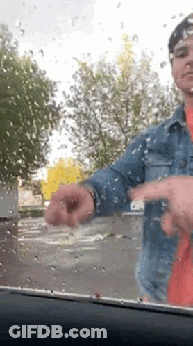 A Man In A Denim Jacket Is Standing In Front Of A Car Windshield With Rain Drops On It . GIF